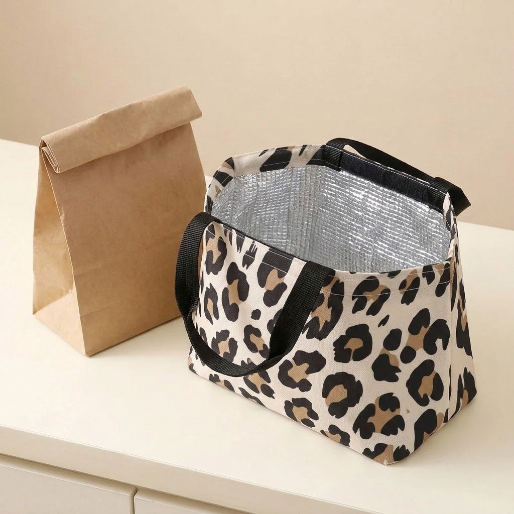 A leopard-print insulated lunch bag with black handles sits on a white table next to a brown paper bag against a beige background.