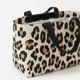 A canvas tote bag with a beige, brown, and black leopard print pattern, featuring black webbed handles and a silver foil-lined interior, shown on a plain white background.