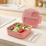 A pink compartment lunch box with a clear lid, shown open on a kitchen counter with food inside and a spoon and chopsticks beside it.
