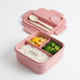 A pink compartment bento box with its lid open, shown on a clean white background. The box contains white rice with black sesame seeds, broccoli, and sliced fruit, and includes a built-in spoon and chopsticks in the lid.
