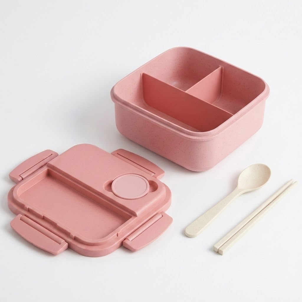 A pink divided bento lunch box shown open with its matching pink lid placed beside it, along with a light beige spoon and chopsticks on a clean white background.