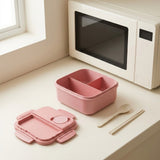 A pink compartment bento box with its matching lid placed beside it on a light countertop, with a microwave and window in the background.