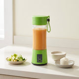 A portable electric blender with a bright green base and lid, filled with an orange smoothie, sits on a white countertop. A small bowl of chopped fruit, a cup of milk, and a plate with powder are arranged nearby in a bright kitchen setting.