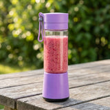 A portable electric blender bottle with a purple lid and base, filled with a pink smoothie, sitting on a weathered wooden table with a blurred green garden background.