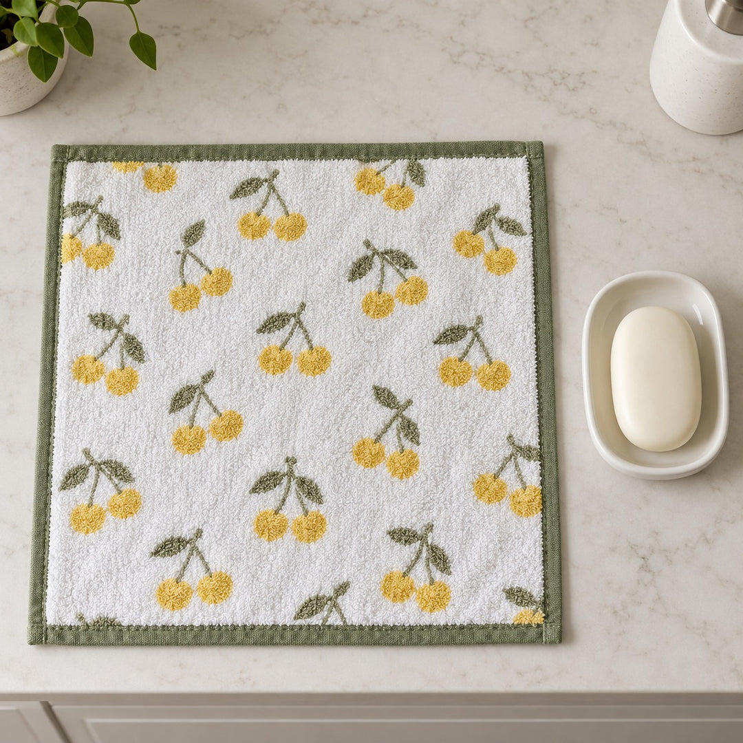 A yellow cherry print cotton washcloth beside a soap dish and bar to show its compact square size.