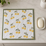 A yellow cherry print cotton washcloth beside a soap dish and bar to show its compact square size.