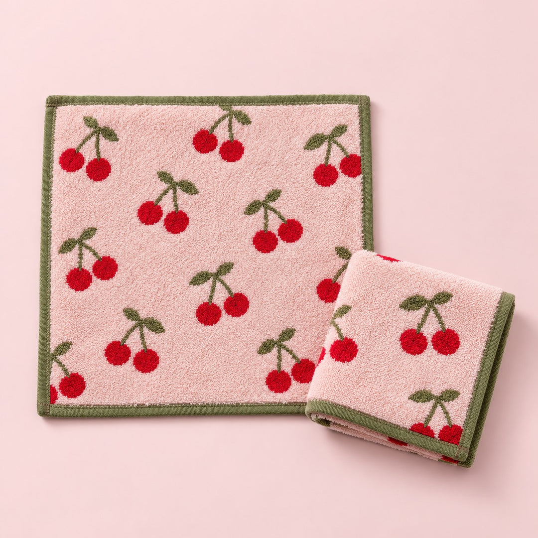 Two red cherry print cotton washcloths shown on a pale pink studio background.