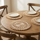 Three round woven placemats in a natural straw color arranged on a round wooden dining table, each styled with a white plate, with wooden chairs visible in a softly lit indoor setting.