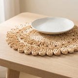 A round woven straw placemat with a scalloped edge sits on a light wood table, styled with a white ceramic plate on top.