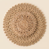 A round, handwoven jute placemat with a detailed concentric circular pattern and fringed edges, displayed flat against a solid cream background.
