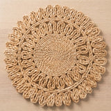 A round woven placemat with a sunburst or rosette pattern, made from natural tan jute or raffia on a light wood background.