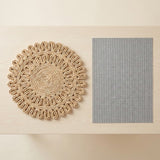 A natural woven round placemat with a scalloped edge sits next to a rectangular gray placemat on a light wood tabletop.