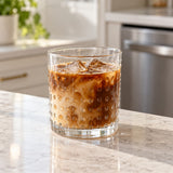 Clear embossed dot drinking glass of iced coffee sits by a dishwasher in a bright kitchen.