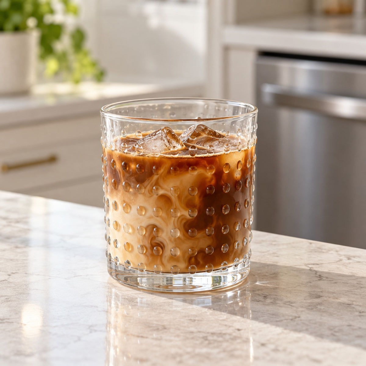Clear embossed dot drinking glass of iced coffee sits by a dishwasher in a bright kitchen.