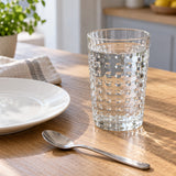 Clear embossed dot drinking glass sits beside a plate and teaspoon on a kitchen table for scale.