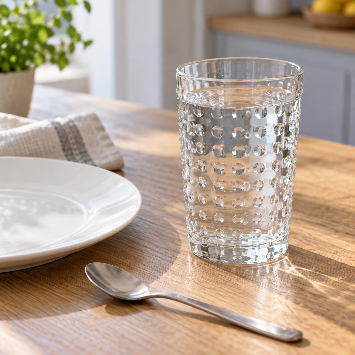 Clear embossed dot drinking glass sits beside a plate and teaspoon on a kitchen table for scale.