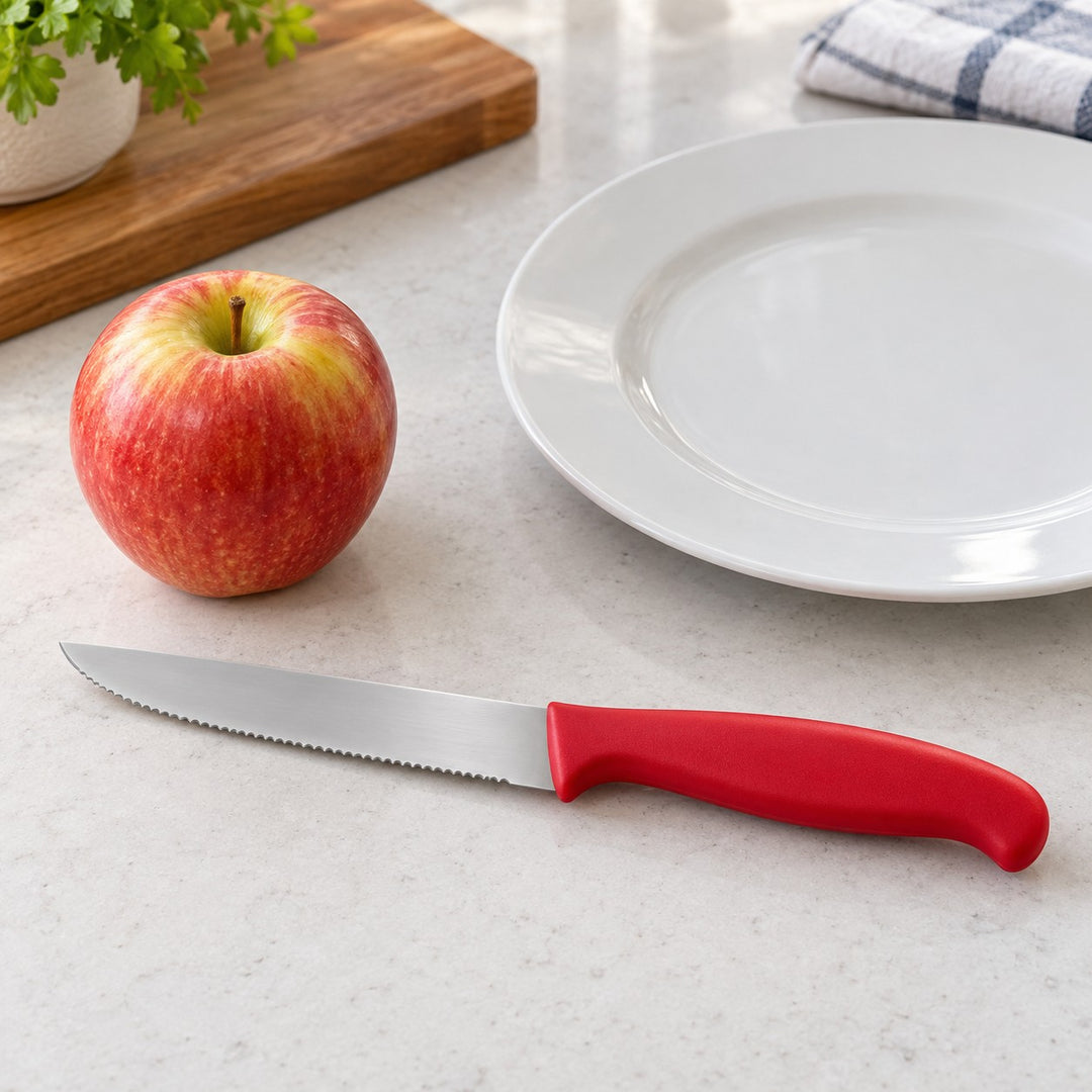 A red Serrated Steak Knife sits beside an apple and dinner plate to show its compact size.