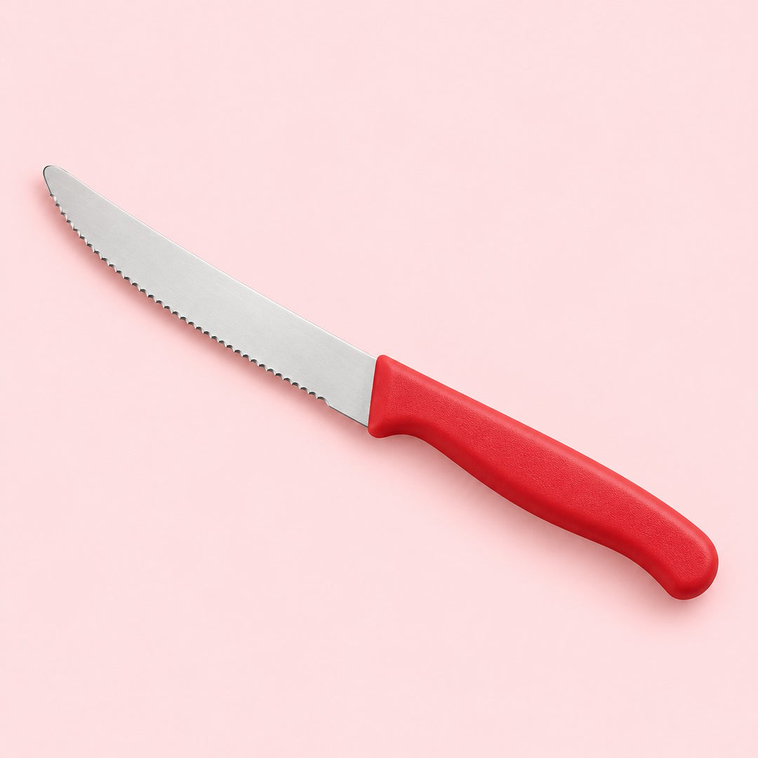 A red Serrated Steak Knife with a rounded stainless steel blade on a pale red studio backdrop.