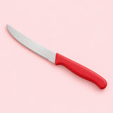 A red Serrated Steak Knife with a rounded stainless steel blade on a pale red studio backdrop.