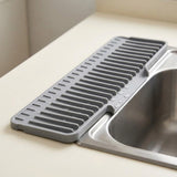 A gray silicone kitchen sink drain guard with a slotted ribbed design, shown installed over the edge of a stainless steel sink on a beige countertop.