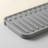 A close-up of a gray silicone dish drying rack with parallel ridges and drainage holes, resting on a light gray surface against a warm off-white background.