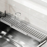 A light gray plastic kitchen sink drain drying rack with a clear glass cup placed on it, set over a stainless steel sink in a kitchen with white tiled walls.