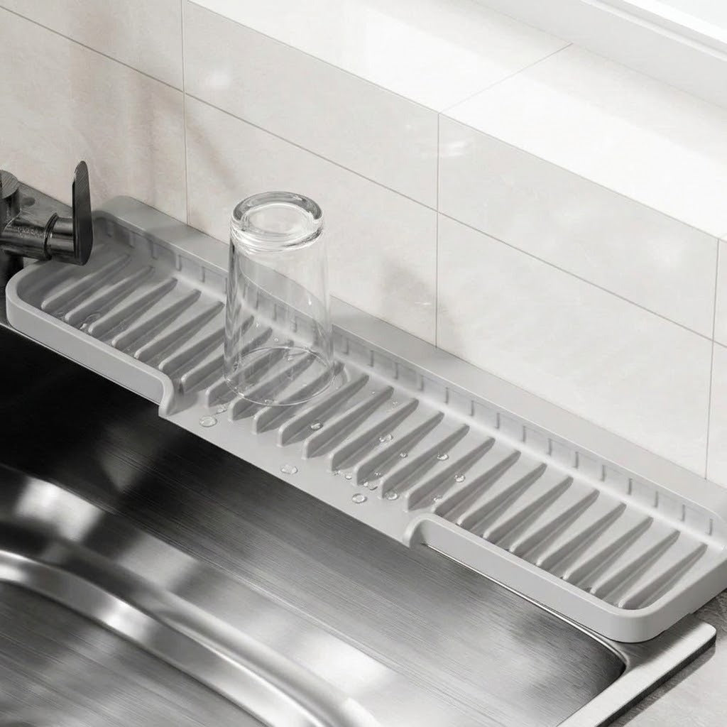 A light gray plastic kitchen sink drain drying rack with a clear glass cup placed on it, set over a stainless steel sink in a kitchen with white tiled walls.