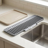 A gray silicone kitchen drying rack placed over a stainless steel sink, with water draining into the basin. A beige tray is set beside it on a light countertop with tiled backsplash.