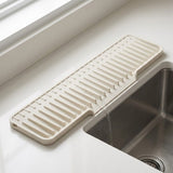 An off-white plastic kitchen sink drain guard with ridges, shown installed over the edge of a stainless steel sink with water draining into it.