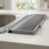 A gray silicone kitchen drying rack with a slotted design, shown resting on a light countertop next to a stainless steel sink in a kitchen setting.