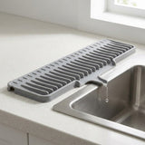 A gray silicone kitchen drying rack with a slotted design, shown resting on a speckled white countertop next to a stainless steel sink with water dripping into it.