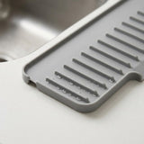 A gray silicone kitchen drying rack with slotted drain channels and water droplets, shown resting on a white countertop near a stainless steel sink.