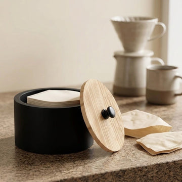 A matte black cylindrical coffee accessory container with a round bamboo lid and small black knob, shown on a countertop with coffee bags and a ceramic pour-over setup in the background.
