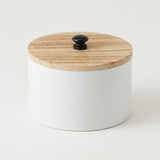 A white cylindrical storage jar with a round light wood lid and a small black knob, photographed on a clean white background.
