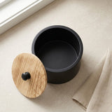 A matte black cylindrical container with a round wooden lid featuring a small black knob, placed on a light beige textured surface near a window.