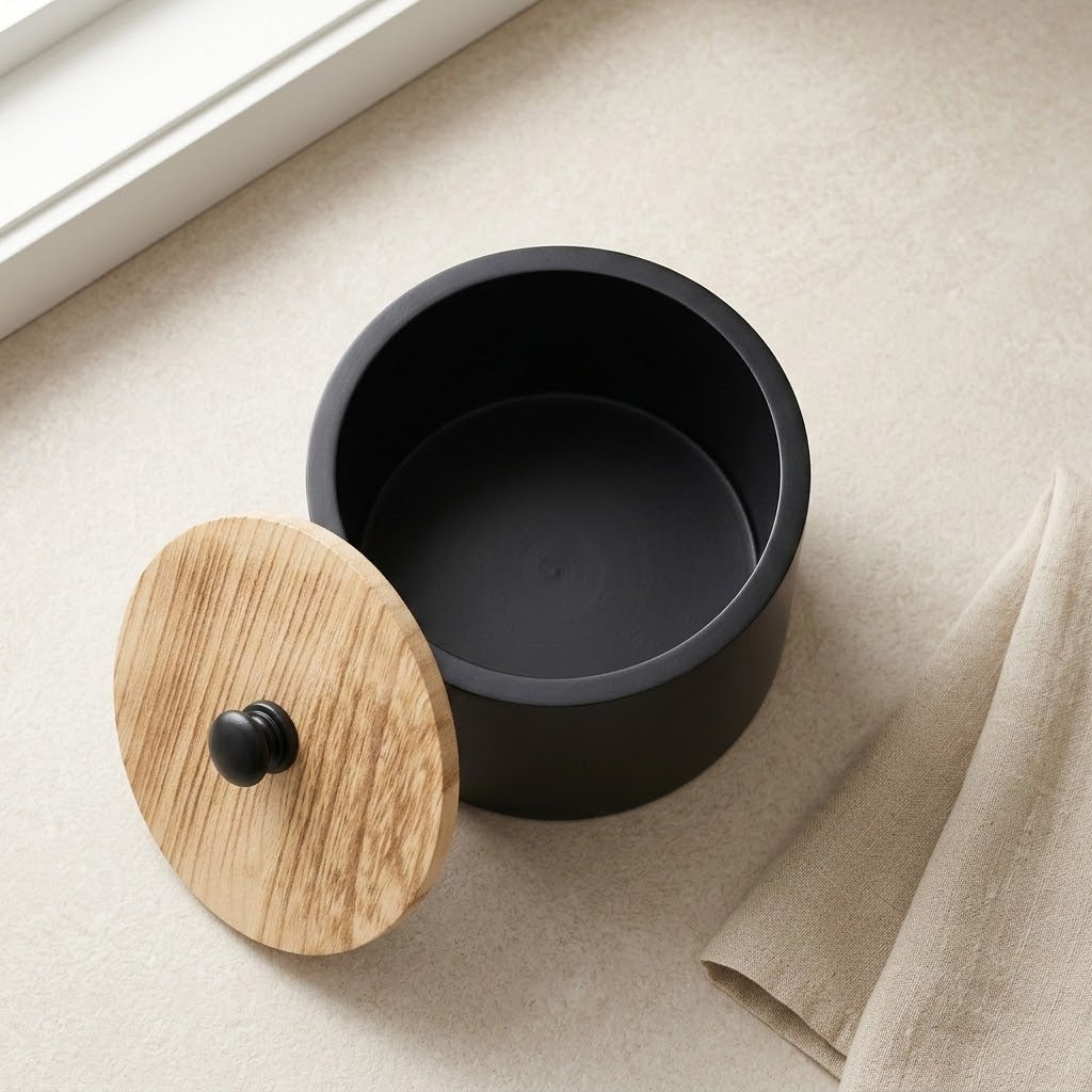 A matte black cylindrical container with a round wooden lid featuring a small black knob, placed on a light beige textured surface near a window.