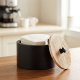 A matte black cylindrical container with a round light wood lid leaning against it, sitting on a wooden table in a softly blurred kitchen setting.