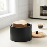 A matte black cylindrical coffee bean jar with a light wood lid and a stack of paper filters inside, set on a kitchen counter near a window with a cup of coffee in the background.