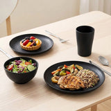 A set of matte black ceramic tableware, including a plate, bowl, and cup, styled on a light wood dining table with food and silverware.