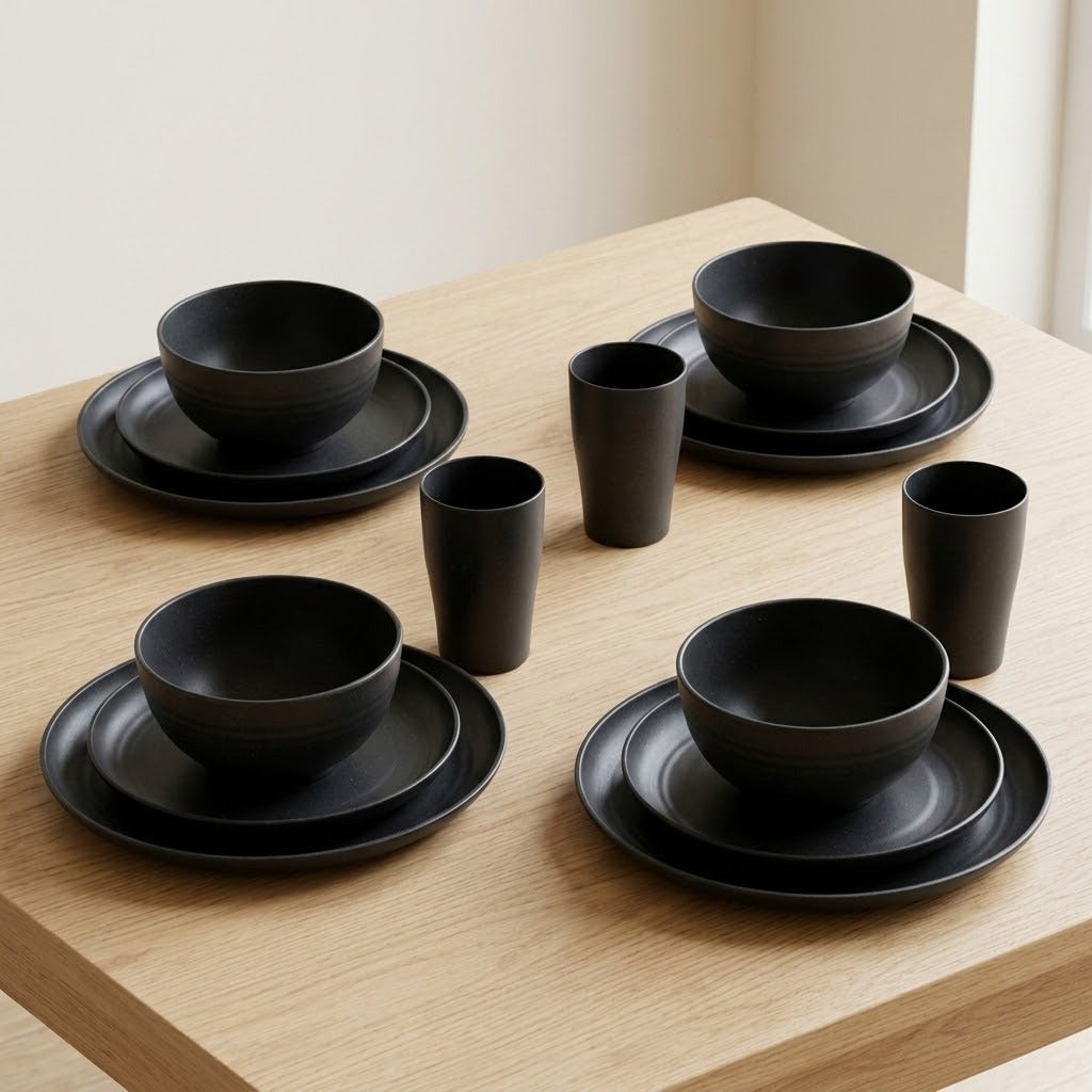 A set of matte black ceramic tableware, including bowls, plates, and cups, arranged on a light wood table against a neutral beige background.