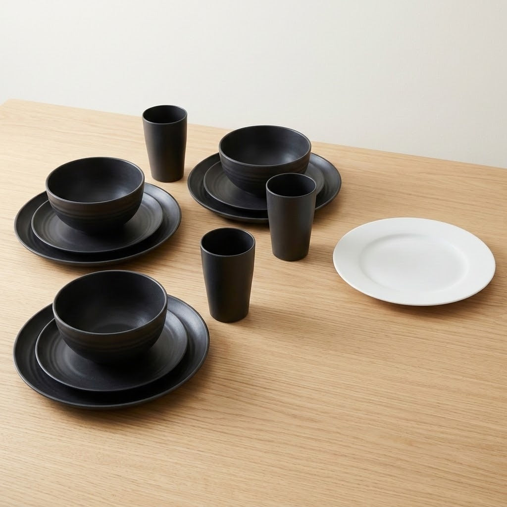A set of matte black dinnerware, including bowls, plates, and cups, arranged on a light wood table next to a single white plate against a neutral background.