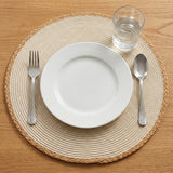 A top-down view of a place setting on a woven round placemat, featuring a white ceramic plate, a silver fork, a silver spoon, and a clear glass of water on a light wood table.