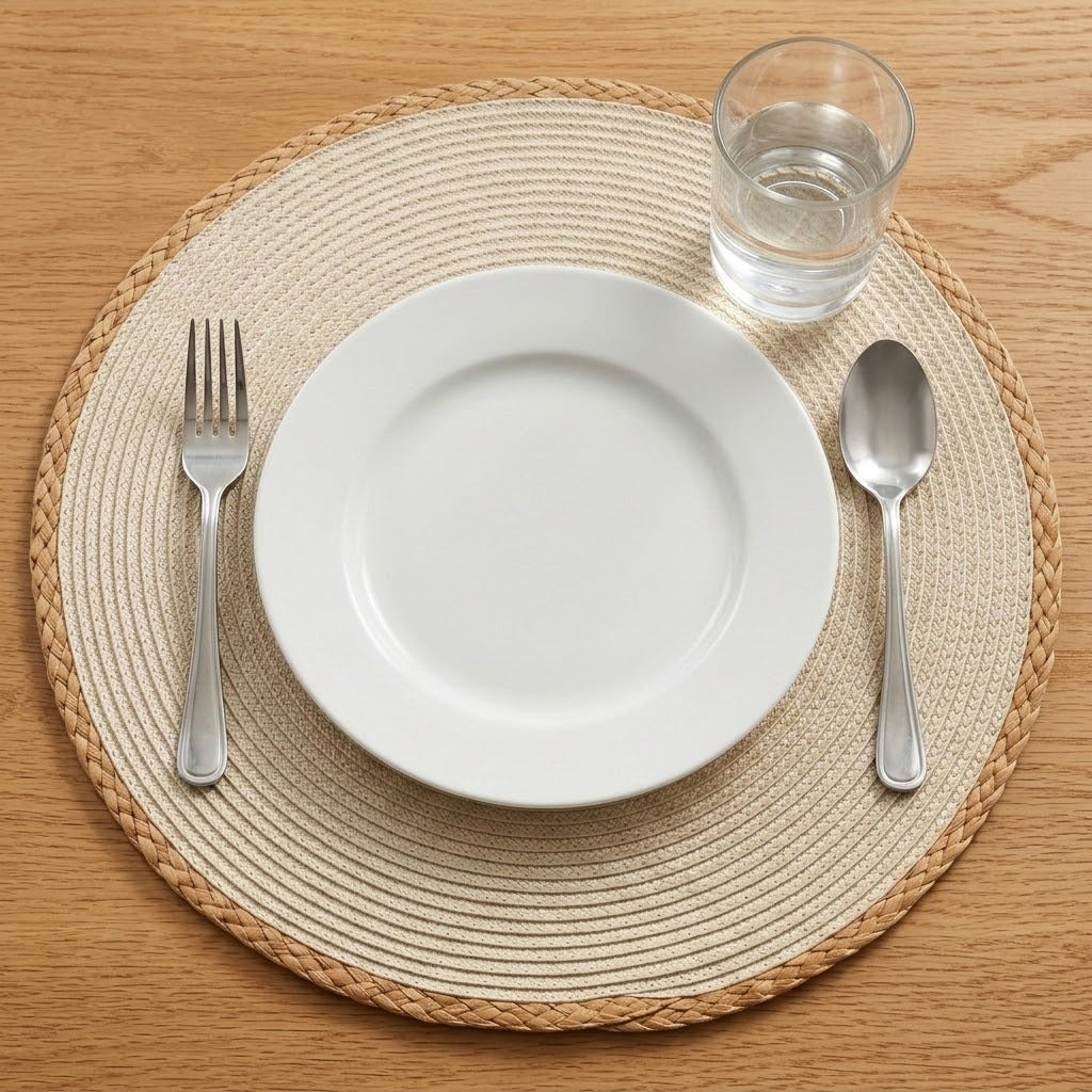 A top-down view of a place setting on a woven round placemat, featuring a white ceramic plate, a silver fork, a silver spoon, and a clear glass of water on a light wood table.