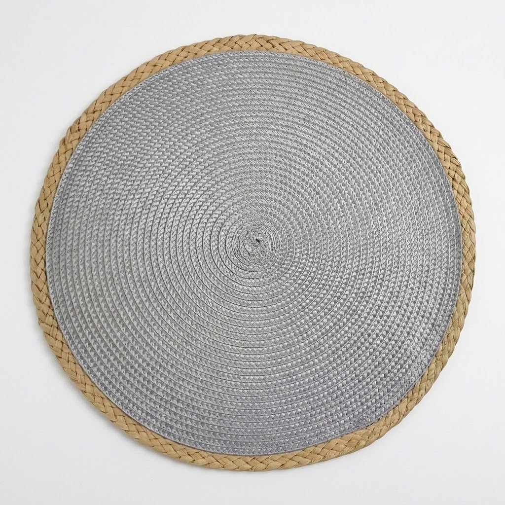 A round woven placemat with a textured silver-gray center and a natural tan braided border, shown from a top-down view on a light gray background.
