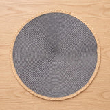 A round woven placemat with a dark gray center and a natural beige braided border, shown from a top-down view on a light wood surface.