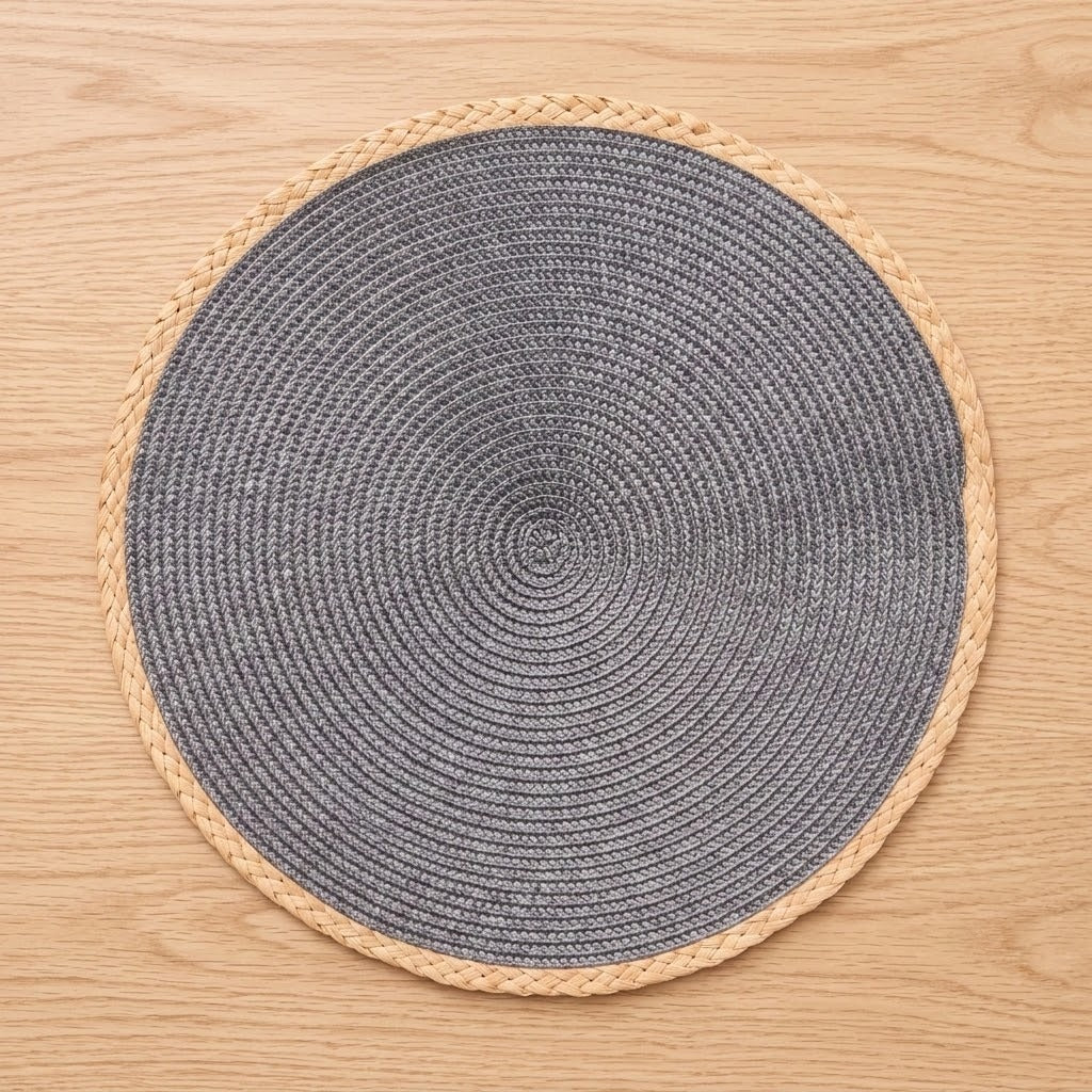 A round woven placemat with a dark gray center and a natural beige braided border, shown from a top-down view on a light wood surface.