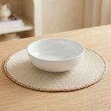 A white ceramic bowl resting on a round woven beige placemat on a light wood table, with a softly blurred indoor background.