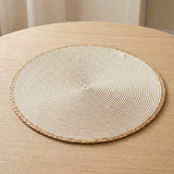 A round woven placemat with a braided edge, shown on a light wood table.
