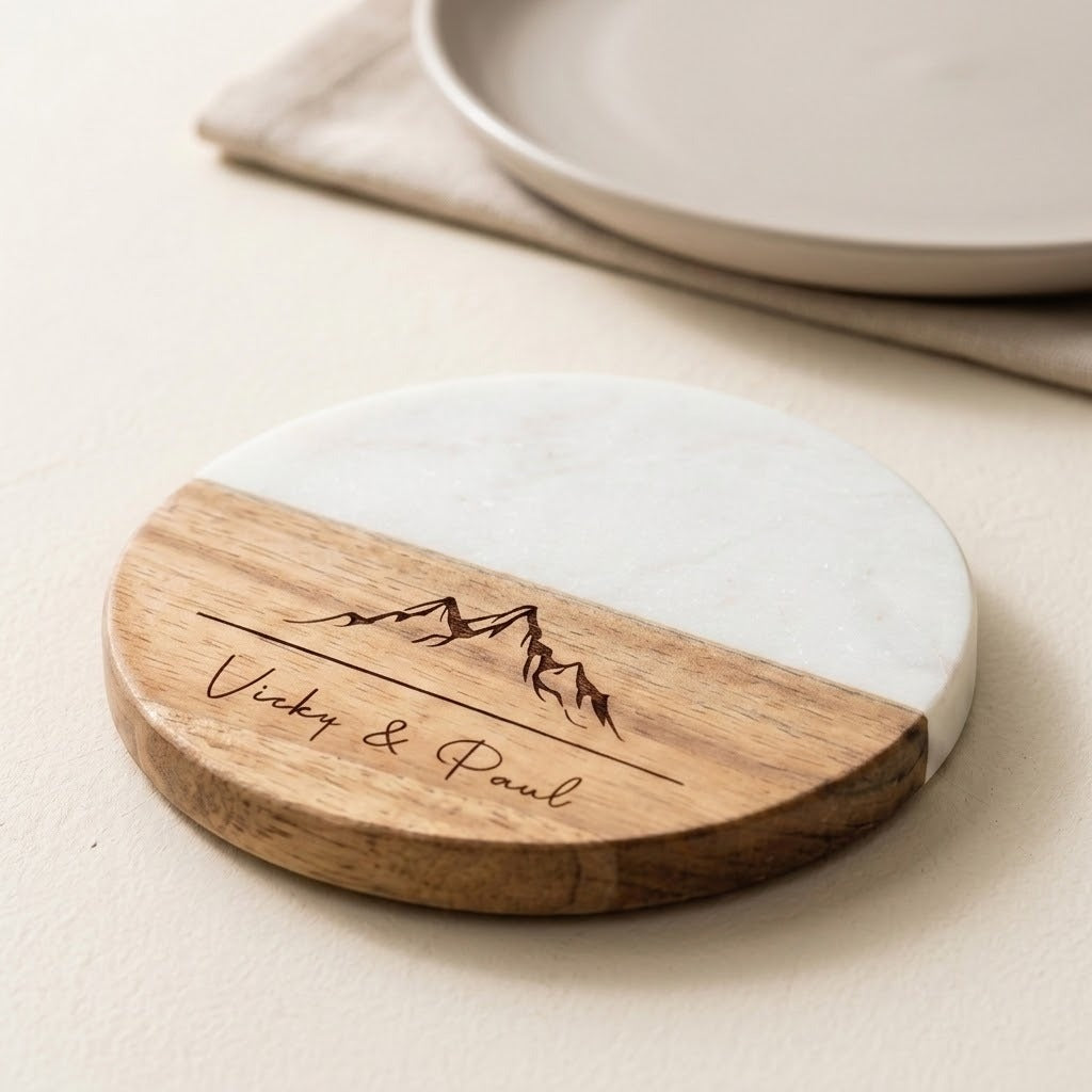 A round wooden and white marble coasters on with engraved mountain graphics and the names 'Vicky & Paul', shown on a light beige tabletop with plates in the background.