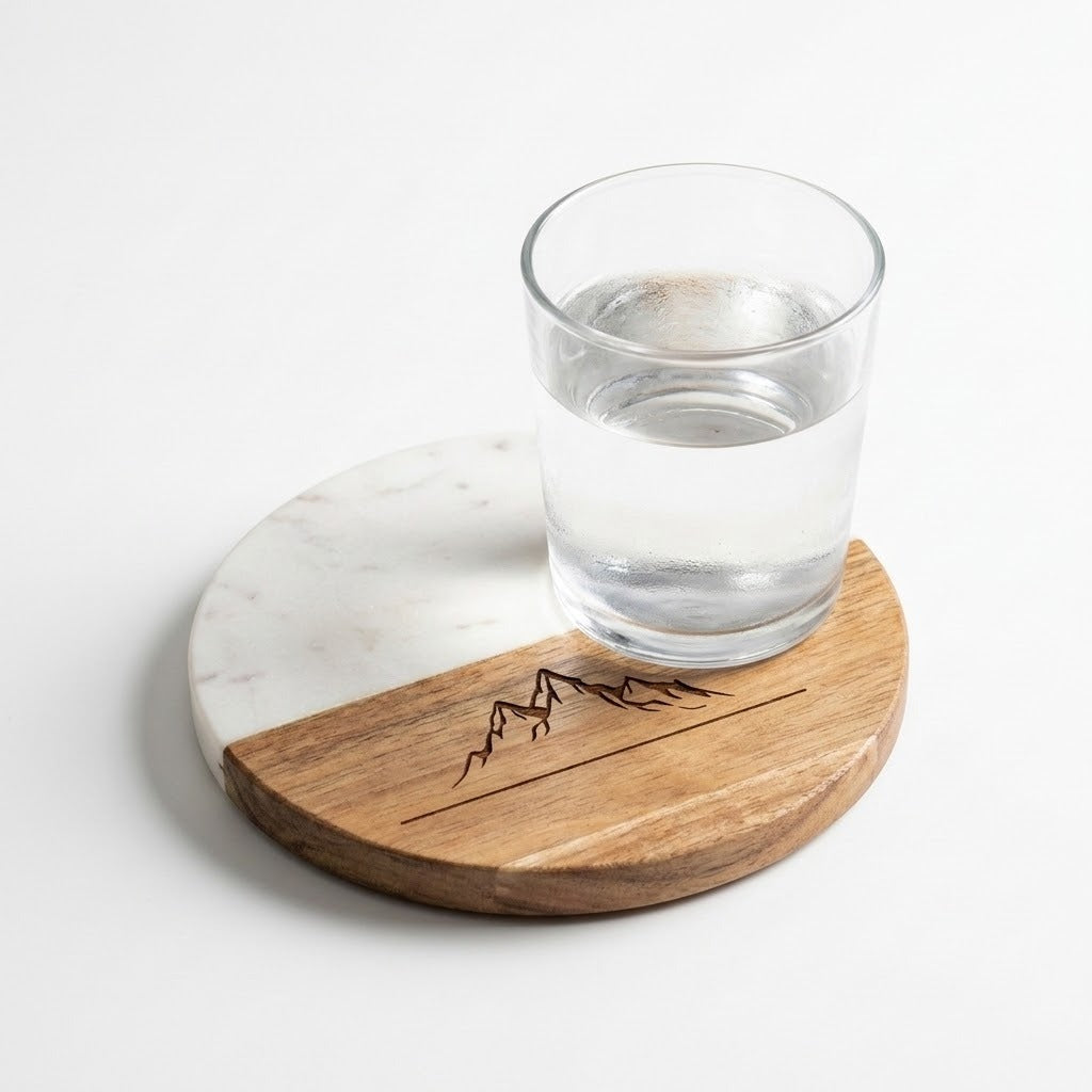 A round wooden and white marble coasters with a dark engraved mountain design, shown with a clear glass of water on a white background.